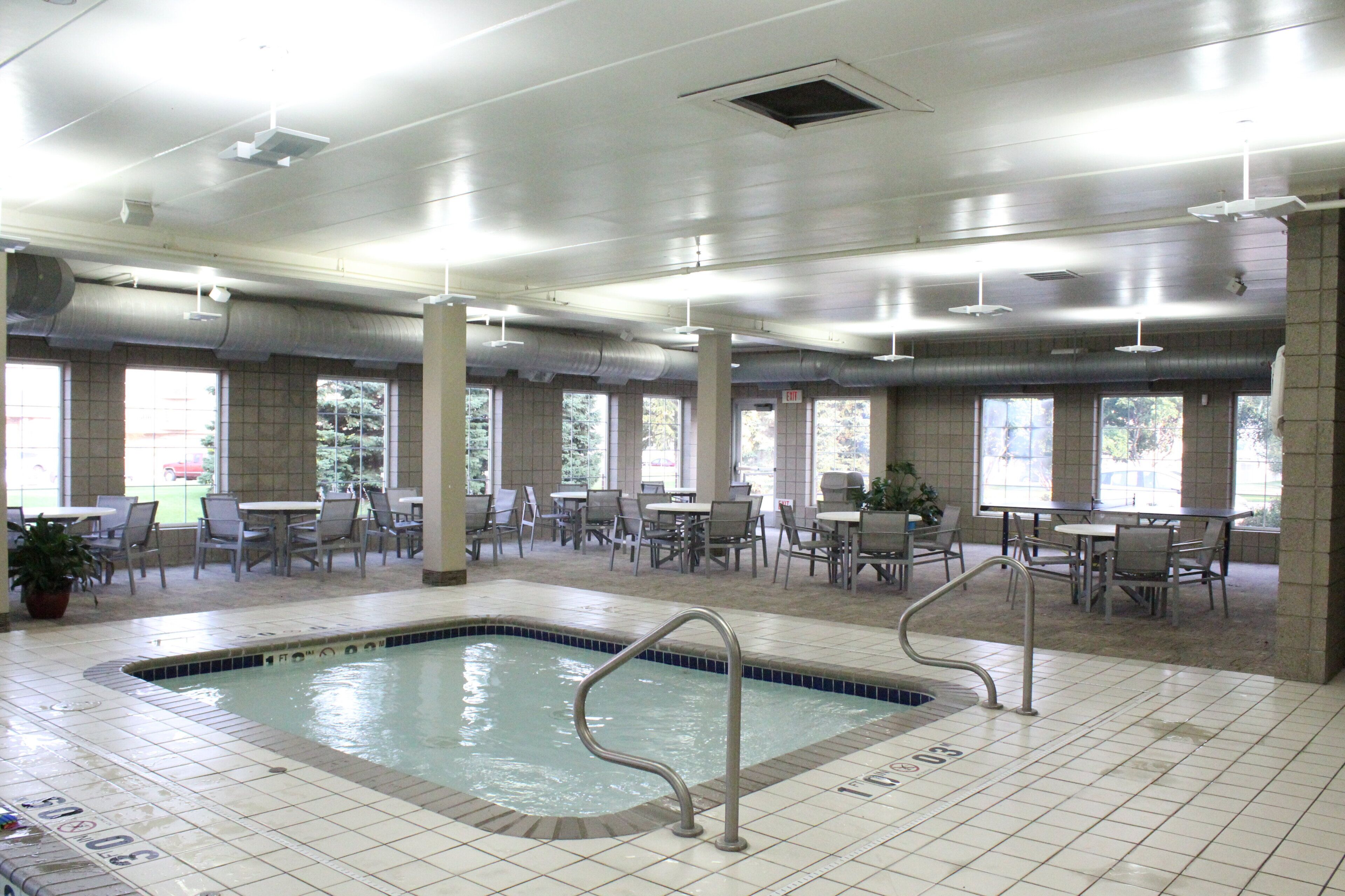 An indoor swimming pool with tiled flooring, surrounded by tables and chairs, and large windows allowing natural light.