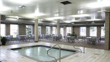 An indoor swimming pool with tiled flooring, surrounded by tables and chairs, and large windows allowing natural light.