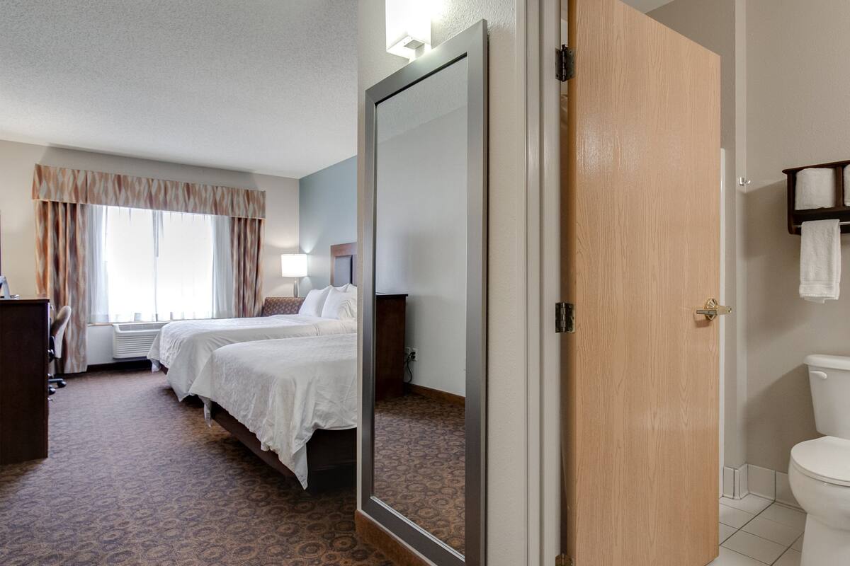 A hotel room with two beds, a bathroom with a toilet and towel rack, and a mirror reflecting part of the room.
