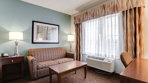 A hotel room with a patterned sofa, a wooden table, a framed picture on the wall, a desk, and a window with curtains. - Holiday Inn Express Hotel & Suites St. Cloud by IHG (St Cloud)