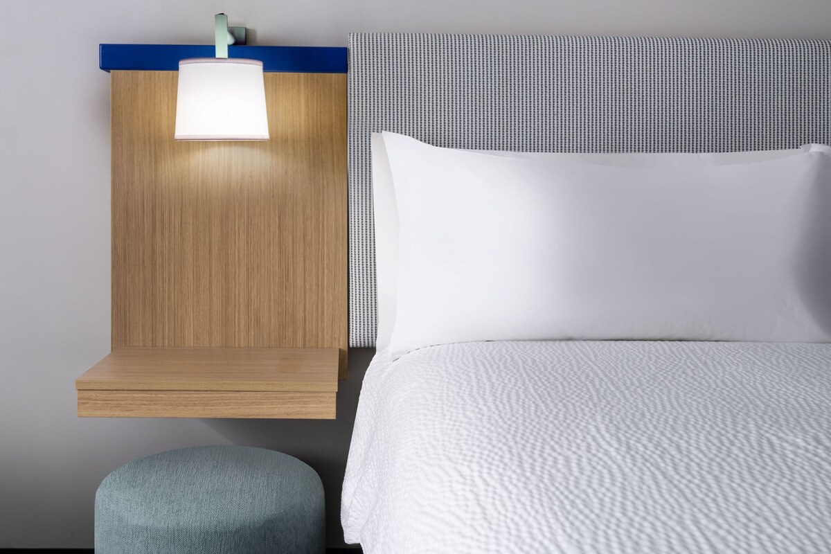 A neatly made bed with a white comforter and a pillow, a bedside table with a lamp, and a small ottoman.