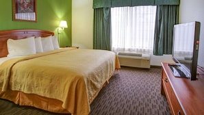 A hotel room with a bed, a desk, a television, a window with curtains, and a wall-mounted air conditioner.