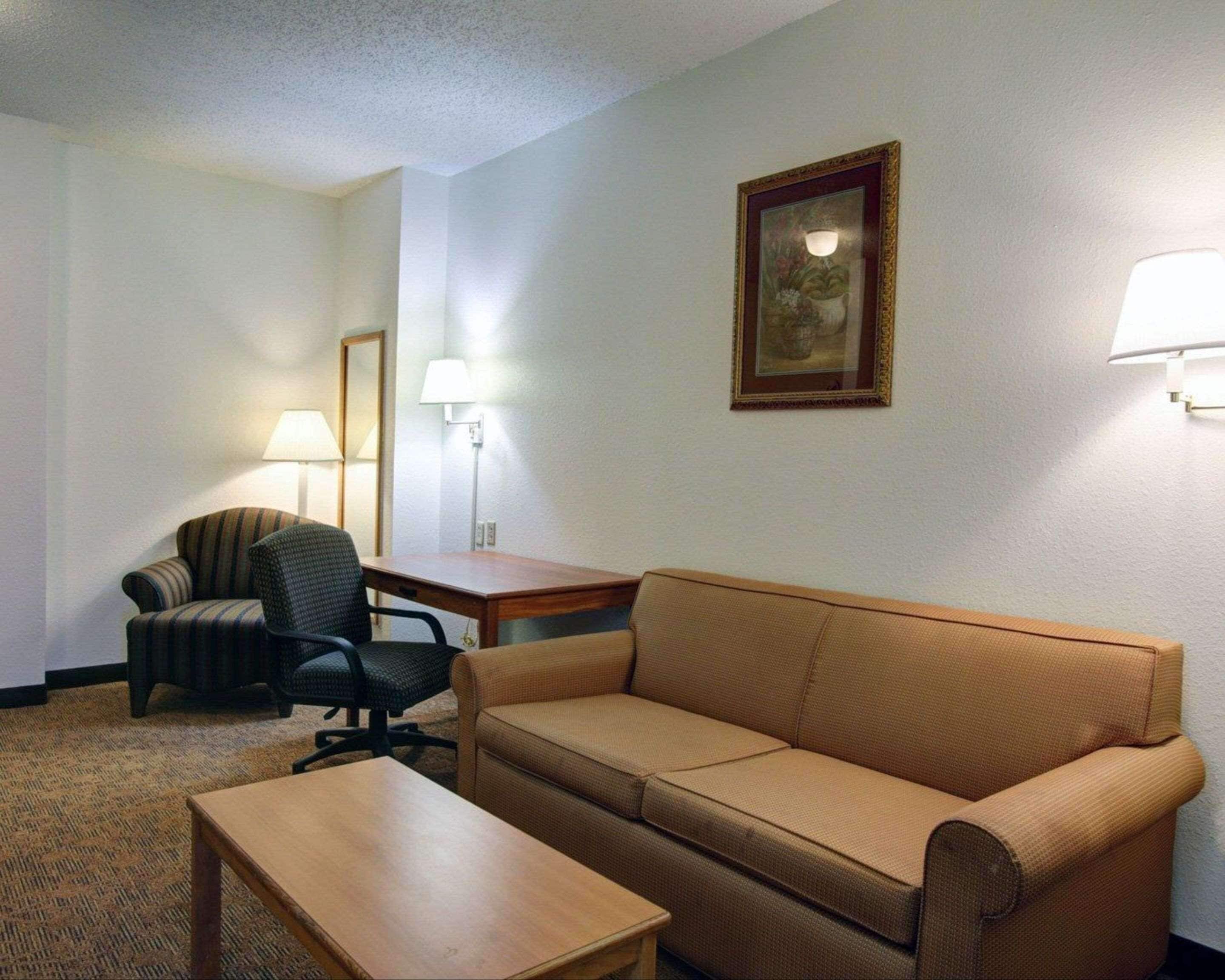 A hotel room with a beige sofa, a wooden table, a striped armchair, and a desk with a lamp.