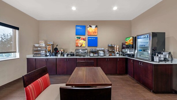 Ein Hotel-Frühstücksbereich mit einem langen Holztisch, gepolsterten Stühlen, einem Kühlschrank und einer Kaffeestation.