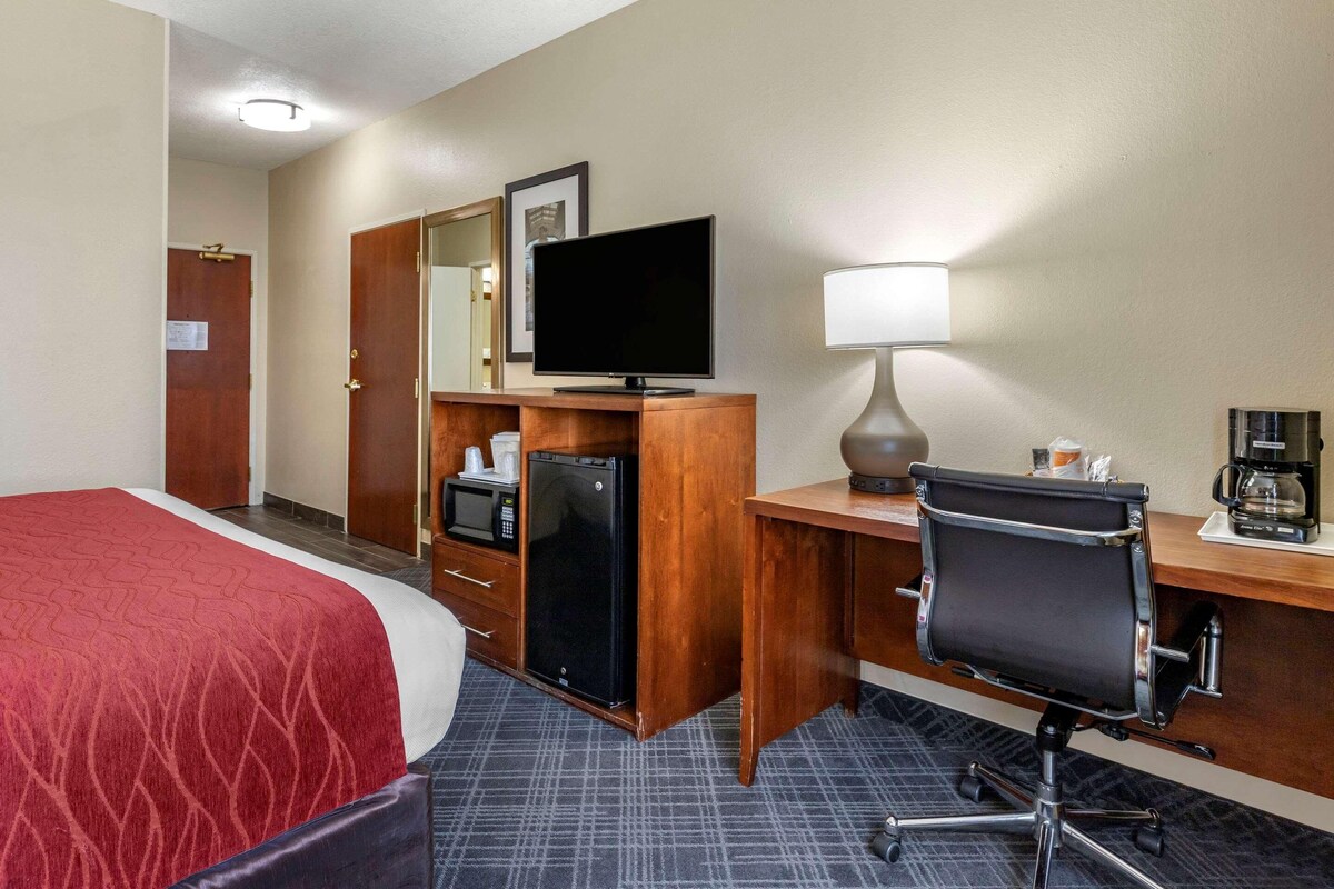 A hotel room with a bed, a desk with a computer, a lamp, a coffee maker, a microwave, and a refrigerator.