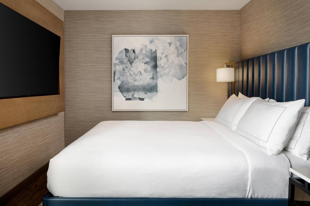 Ett hotellrum med en stor säng, en platt-TV som är monterad på väggen, en modern, abstrakt målning och en blå sänggavel med en sänglampa.