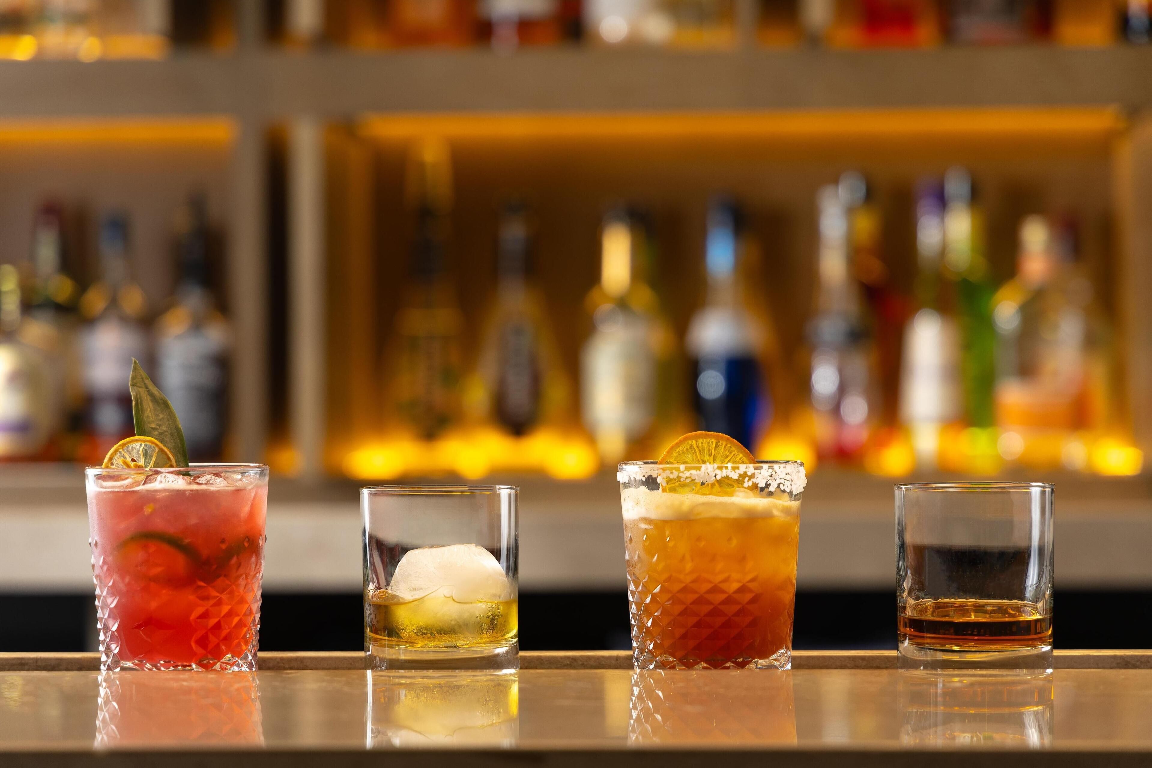 Four glasses of different beverages on a bar counter with a blurred background of bottles.