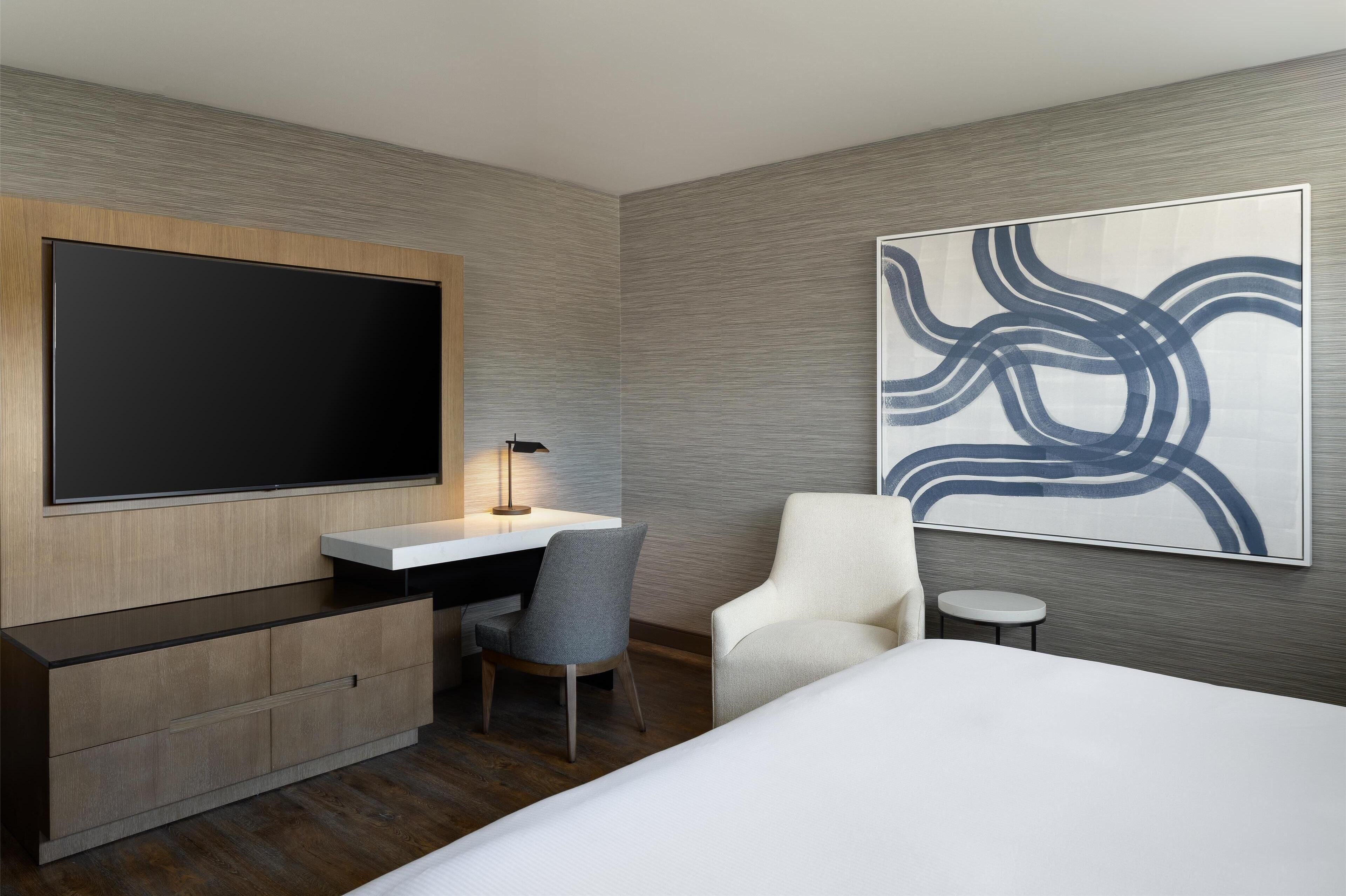 Una habitación de hotel moderna con una televisión de pantalla plana grande, un escritorio con lámpara, una silla gris, un sillón blanco y un cuadro abstracto grande en la pared.