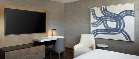 A modern hotel room with a large flat-screen TV, a desk with a lamp, a gray chair, a white armchair, and a large abstract painting on the wall.