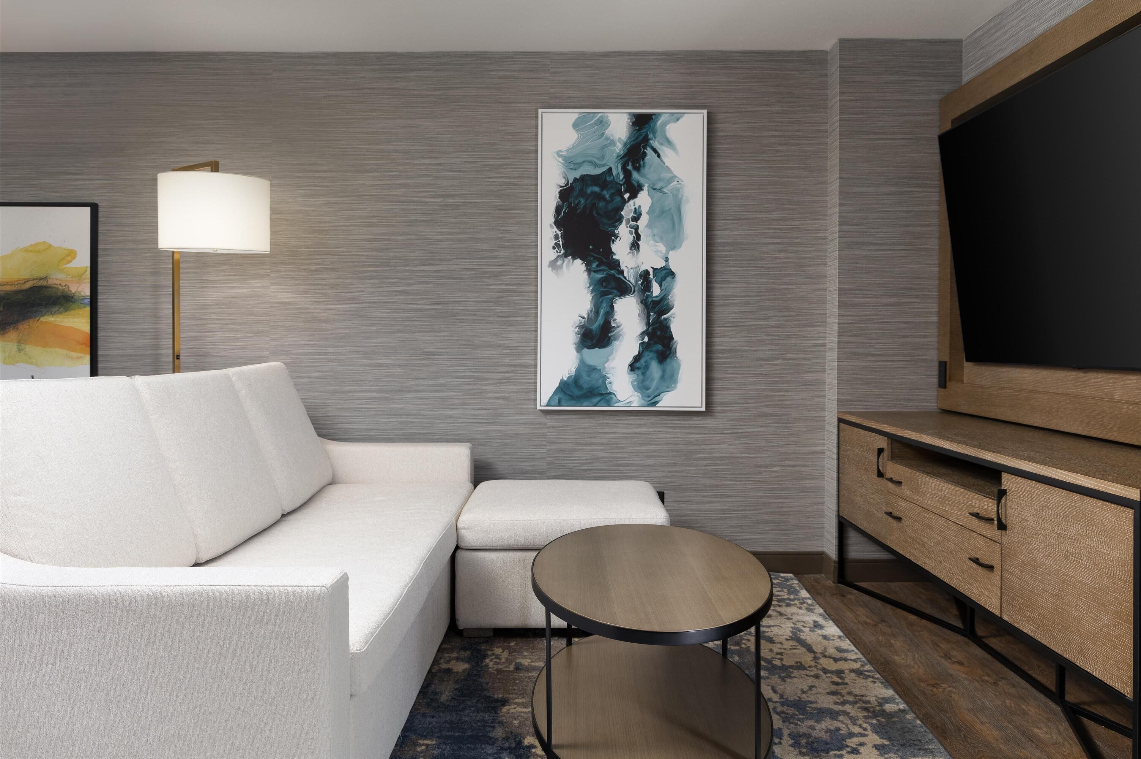 A modern living room with a white sofa, a small round table, a large abstract painting, a floor lamp, and a TV on a wooden cabinet.