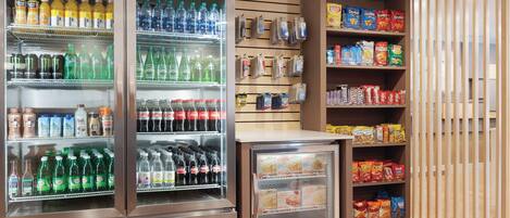 A self-service beverage and snack area with refrigerated drink dispensers, shelves stocked with various beverages and snacks, and a small refrigerator.