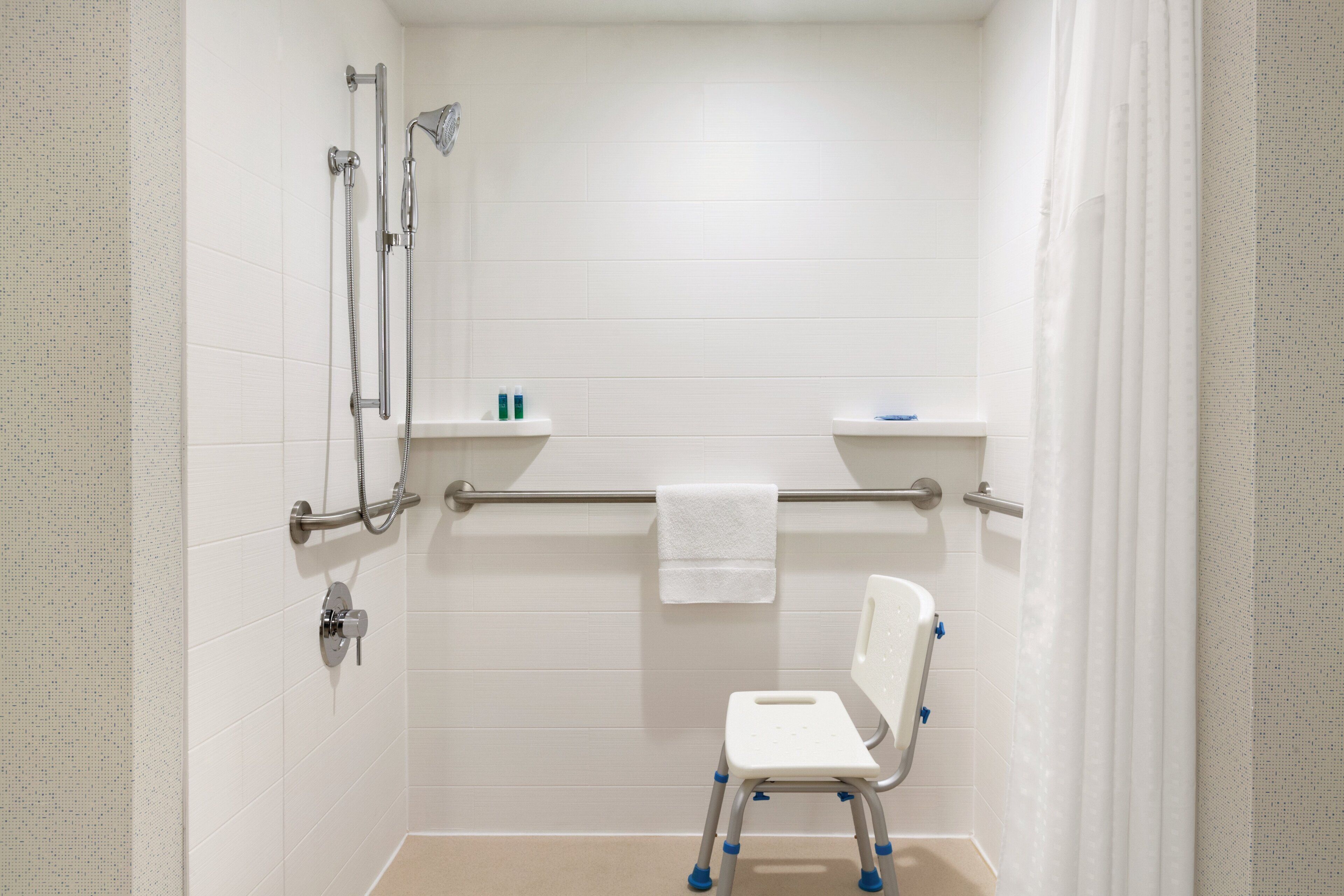 Une salle de bain avec des murs recouverts de carreaux blancs, une douche avec un pommeau de douche amovible, une barre d’appui, des toilettes blanches et un abattant blanc avec des supports bleus.