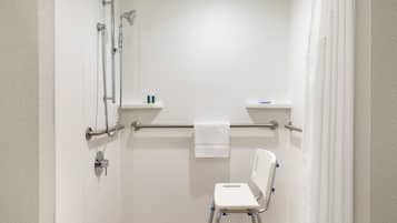 Une salle de bain avec des murs recouverts de carreaux blancs, une douche avec un pommeau de douche amovible, une barre d’appui, des toilettes blanches et un abattant blanc avec des supports bleus.