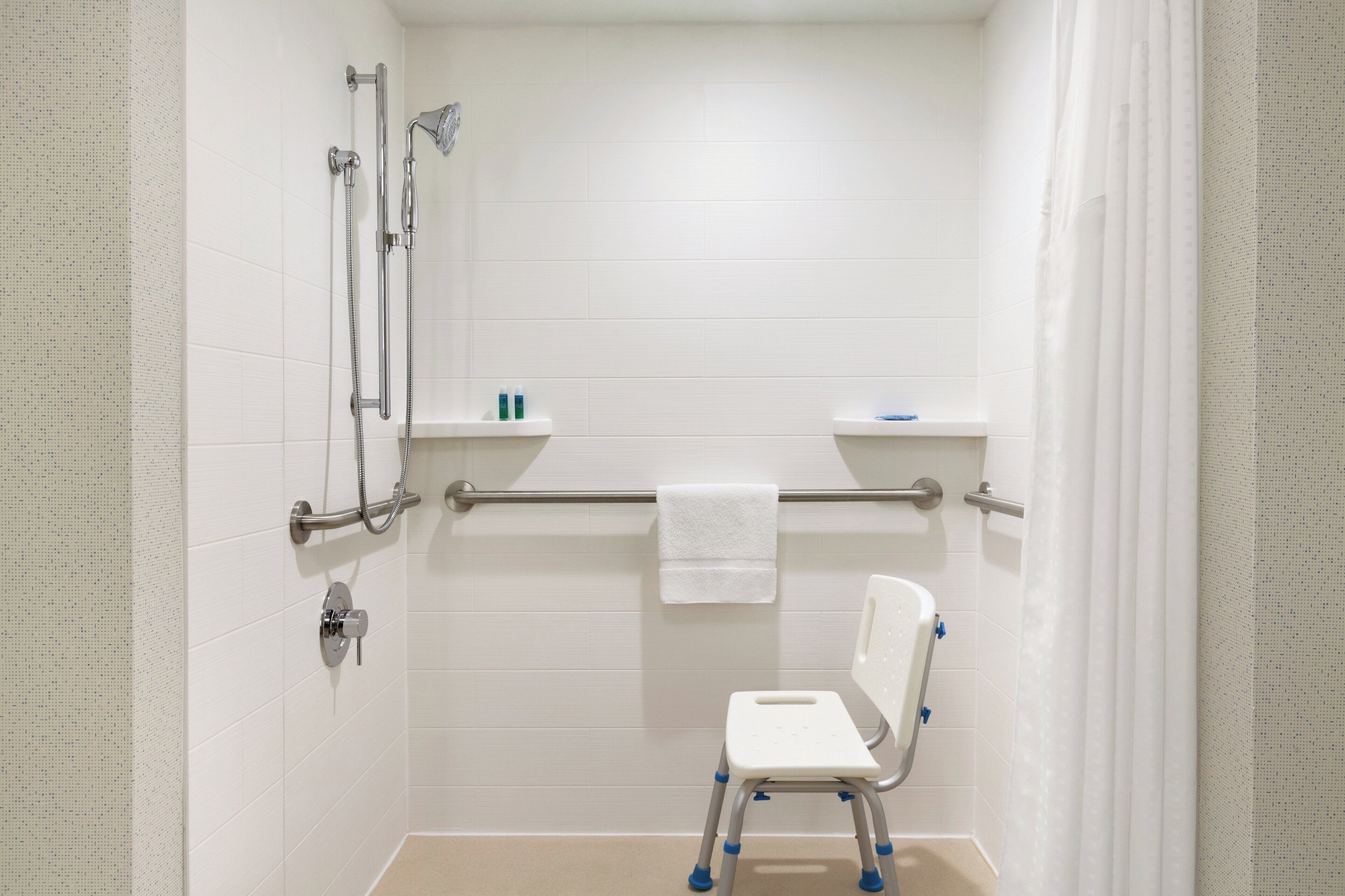 A bathroom with white tiled walls, a shower with a handheld showerhead, a grab bar, and a white step stool.