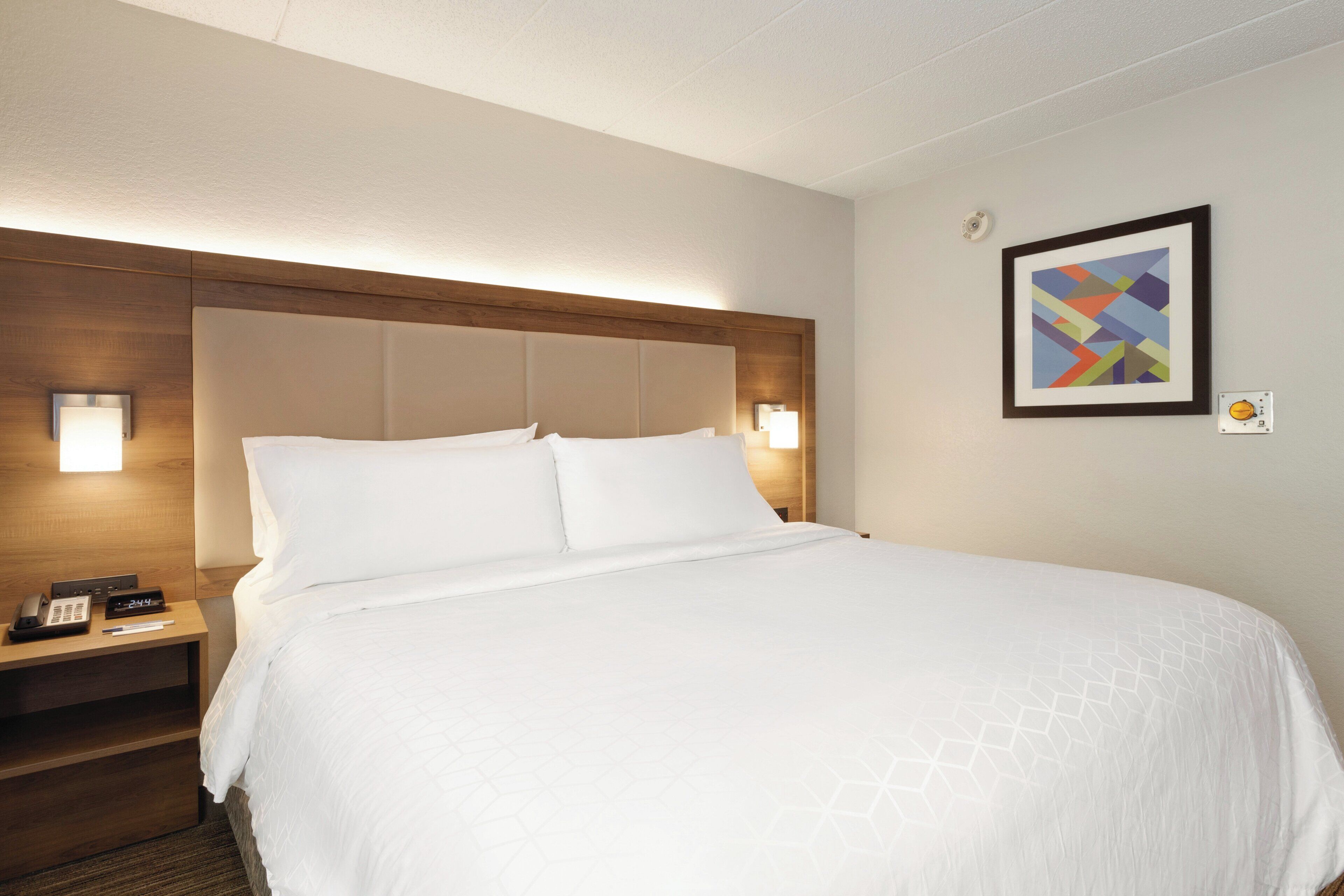 Habitación de hotel con cama, lámparas de noche, mesita con teléfono e imagen enmarcada en la pared.