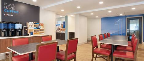 Un comedor moderno con sillas rojas y una mesa larga, una estación de café con varios dispensadores y una sección de panadería al fondo.