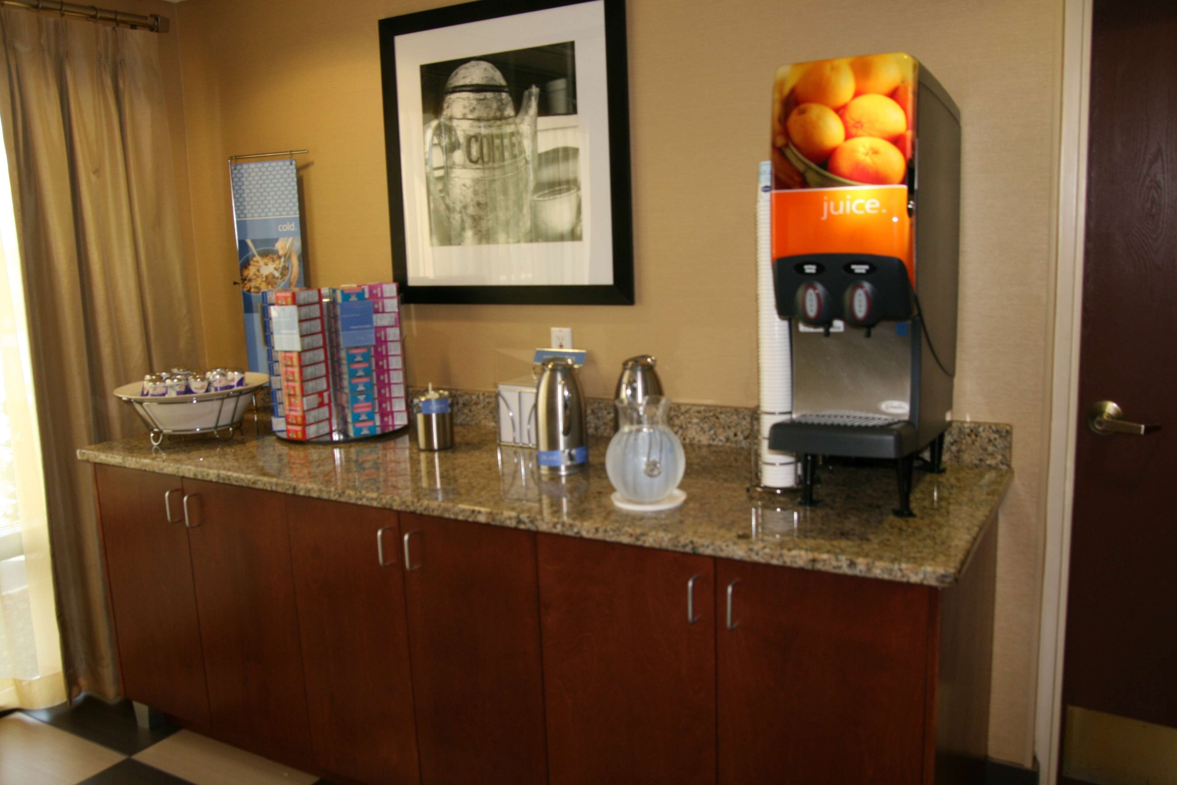 Uma área de recepção de hotel com uma máquina de café, um dispensador de suco, um quadro emoldurado e uma cesta de frutas.