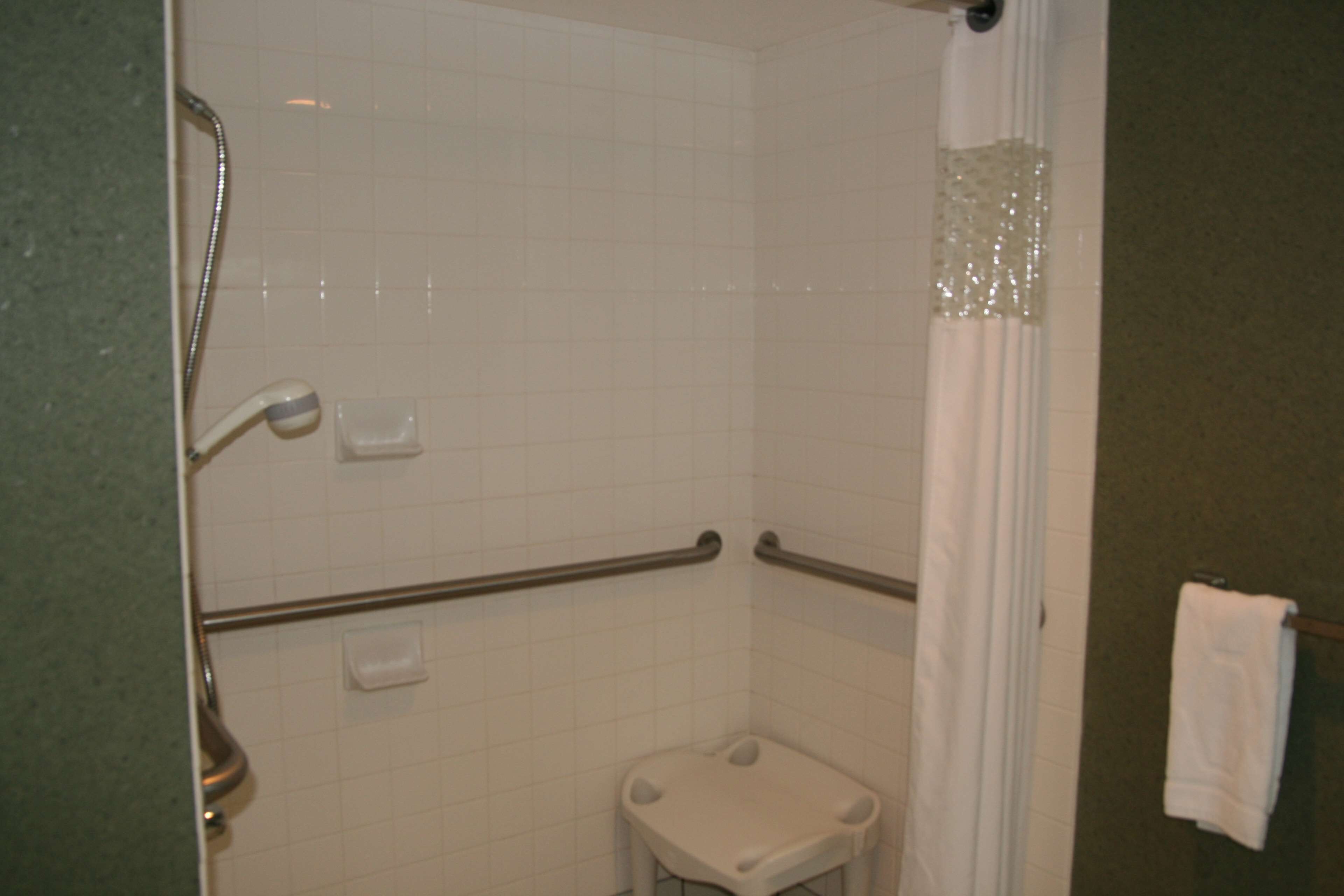 Un baño con paredes de azulejos blancos, una ducha con puerta de cristal, un asidero y un taburete pequeño blanco.