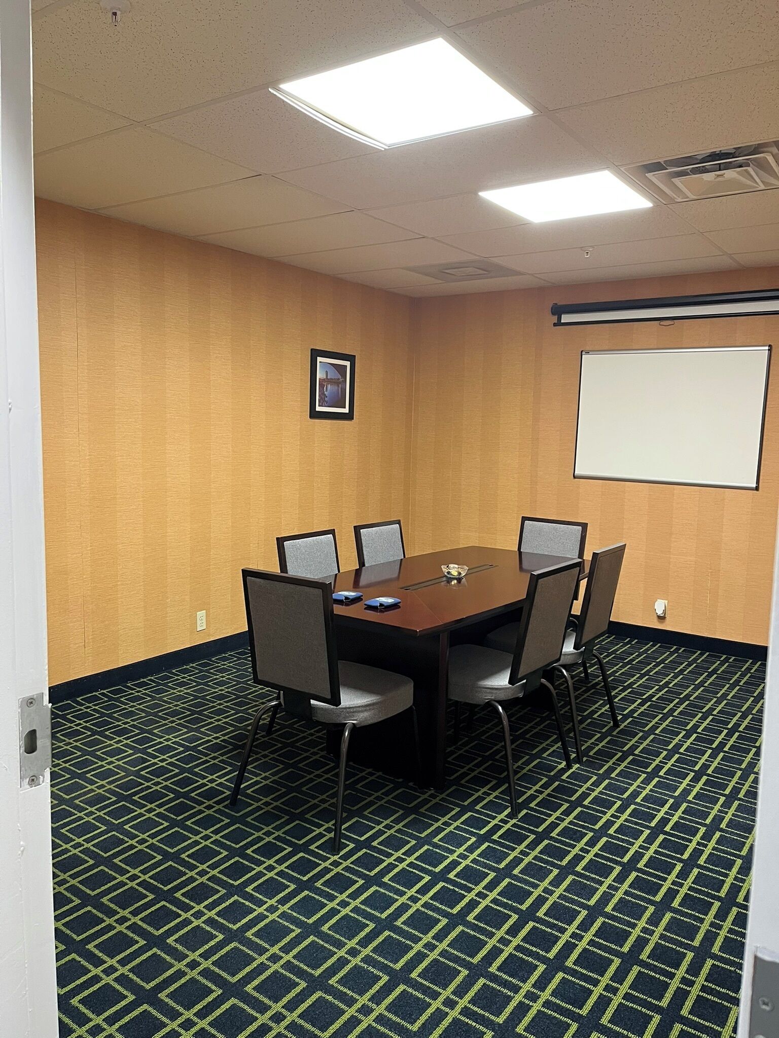 Un salón de conferencias con una mesa rectangular, seis sillas, una pantalla de proyector y un cuadro en la pared.