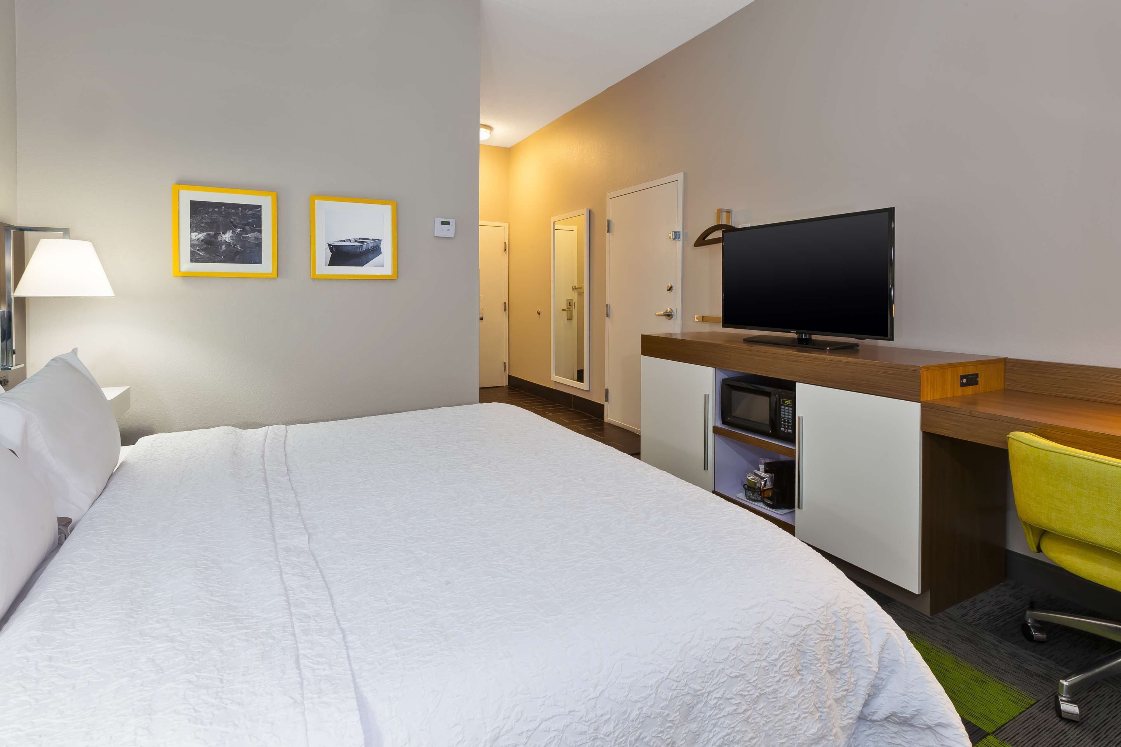 A hotel room with a large bed, a desk with a television, a chair, and two framed pictures on the wall.