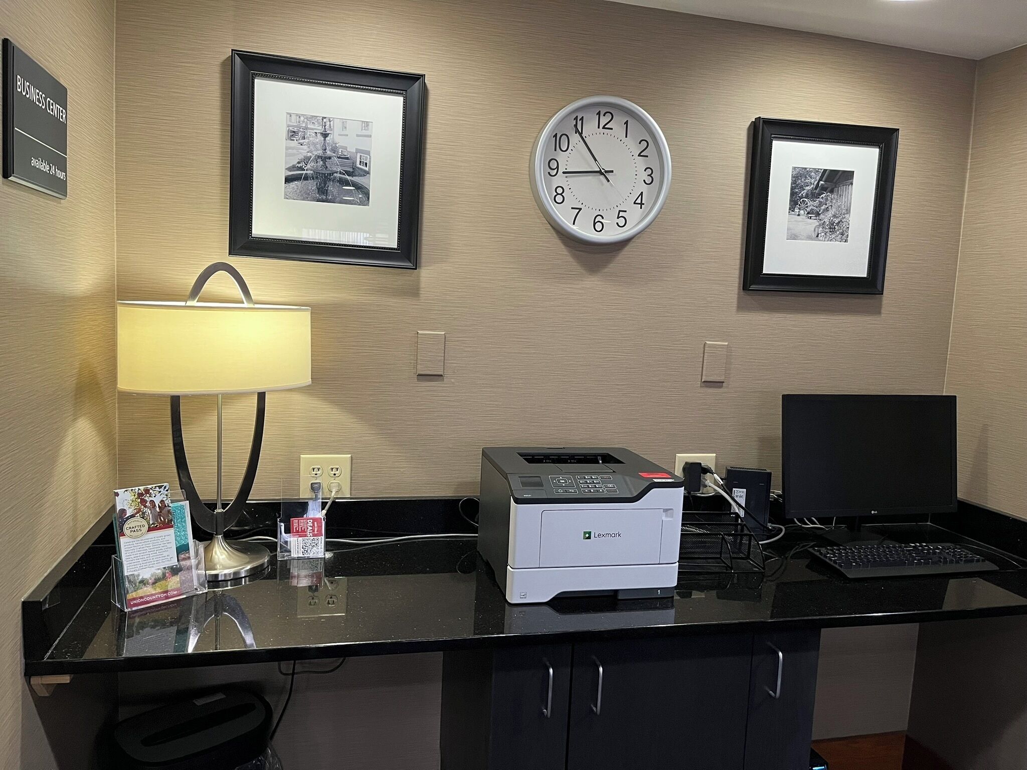 A hotel reception desk with a clock, framed pictures, a lamp, and a Lexmark printer.