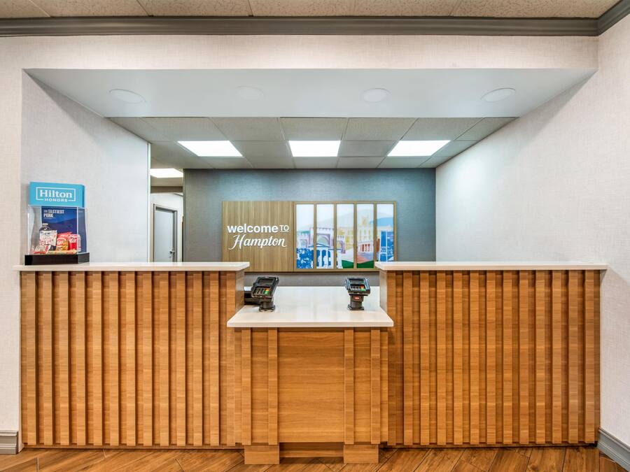 Ein Empfangsschalter mit Holzoptik, ein Schild mit der Aufschrift 'Welcome to Hampton' und ein Hilton Honors-Schild.
