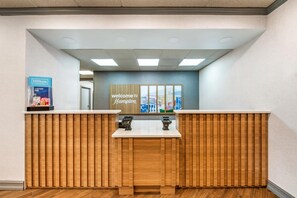 Un mostrador de recepción con detalles de madera, un cartel que dice 'Welcome to Hampton' y el logo de Hilton Honors.