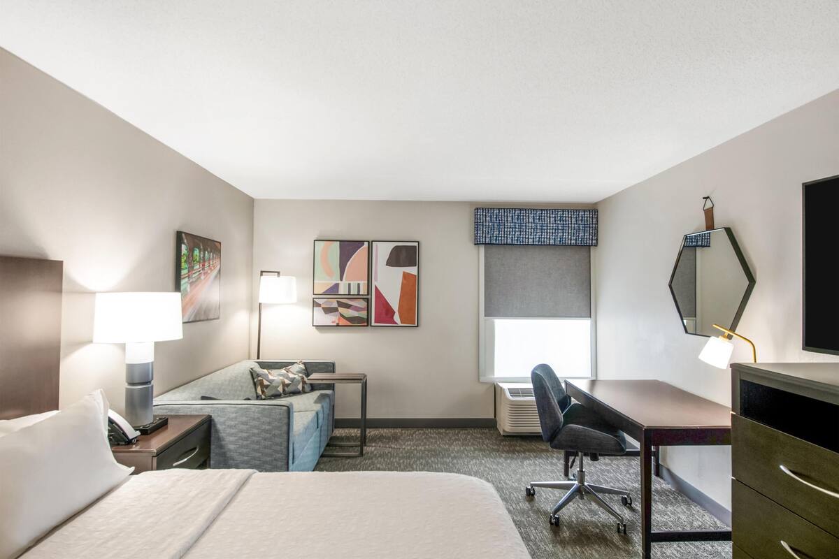 Een hotelkamer met een bed, bureau, stoel, lamp, airconditioning en kunst aan de muur.