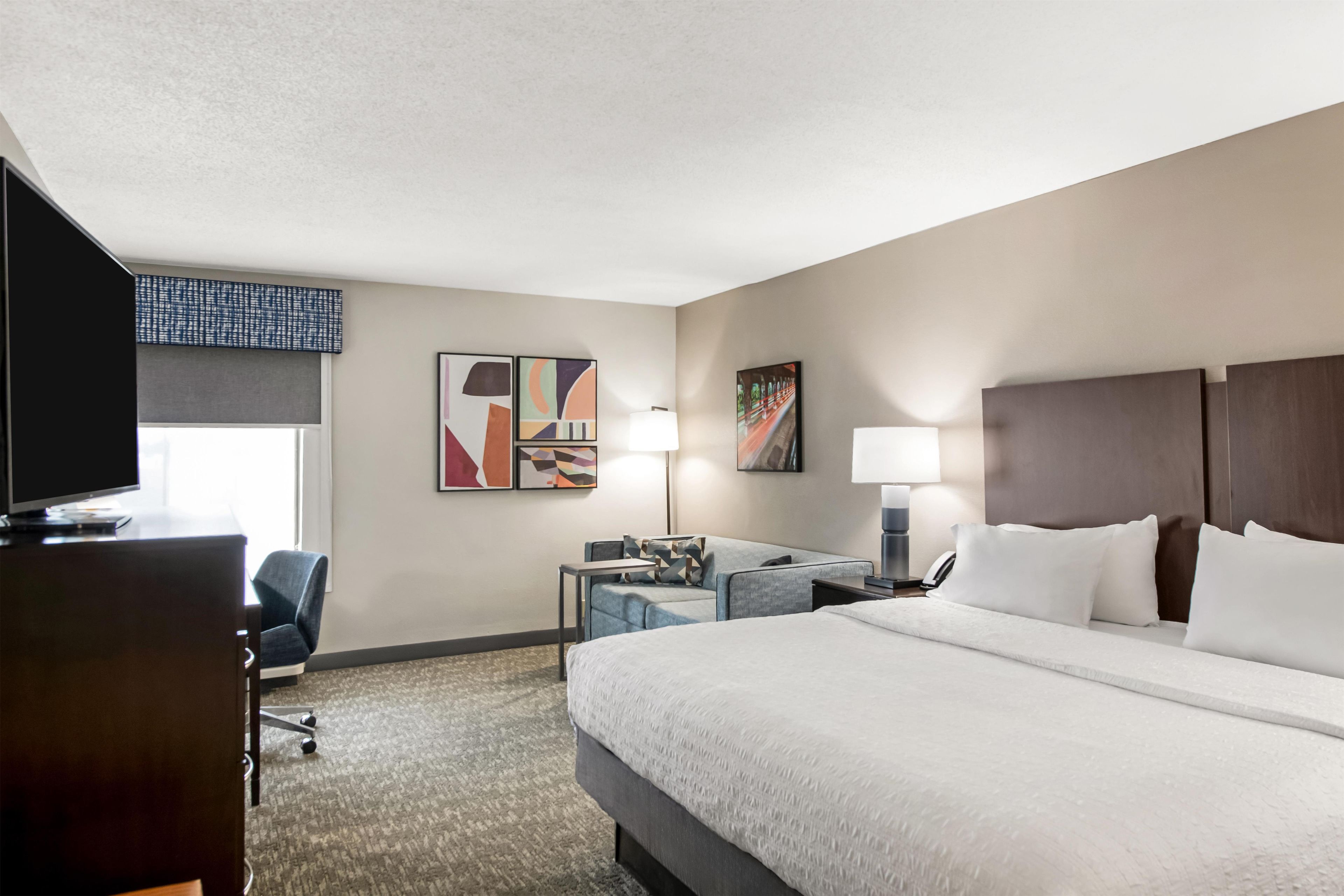 A hotel room with a large bed, a desk, a chair, a television, and artwork on the walls.