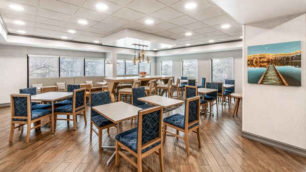 A spacious dining area with wooden tables and blue upholstered chairs, large windows, and a painting on the wall. - Hampton Inn Milledgeville (Milledgeville)