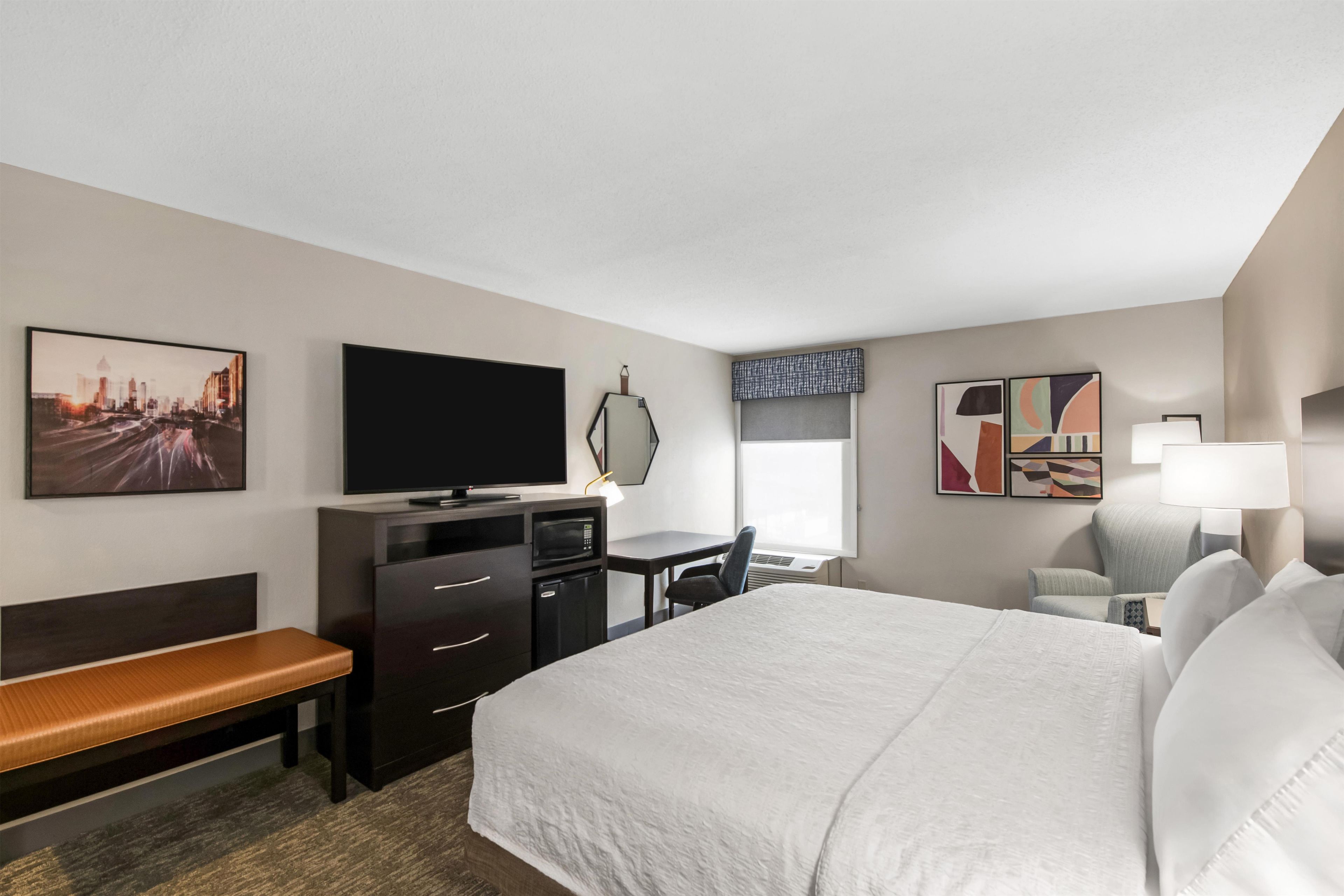 A hotel room with a bed, a desk, a television, a bench, and artwork on the walls.