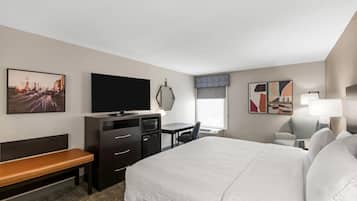 A hotel room with a bed, a desk, a television, a bench, and artwork on the walls.