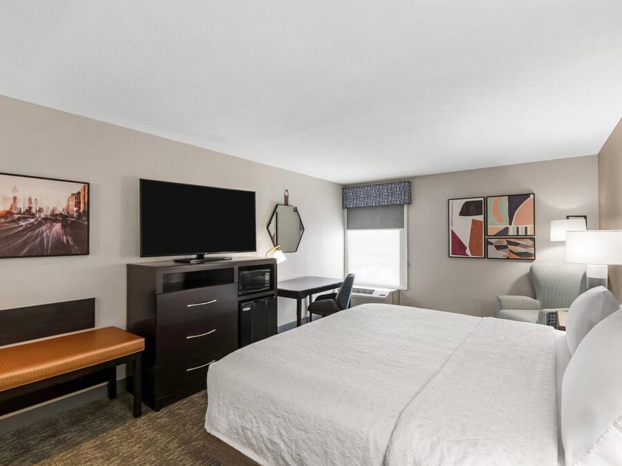 A hotel room with a bed, a desk, a television, a bench, and artwork on the walls.