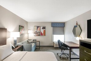 Habitación de hotel con cama, escritorio, silla, lámpara de escritorio, televisor montado en la pared y ventana con persianas.