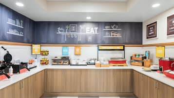 Un área de desayuno con una pared de pizarra que muestra dibujos y texto relacionados con comida, una máquina de café y varios alimentos para el desayuno sobre la encimera.