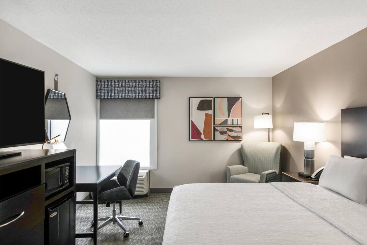 Een hotelkamer met een bed, bureau, stoel, televisie, kunst en lampen.