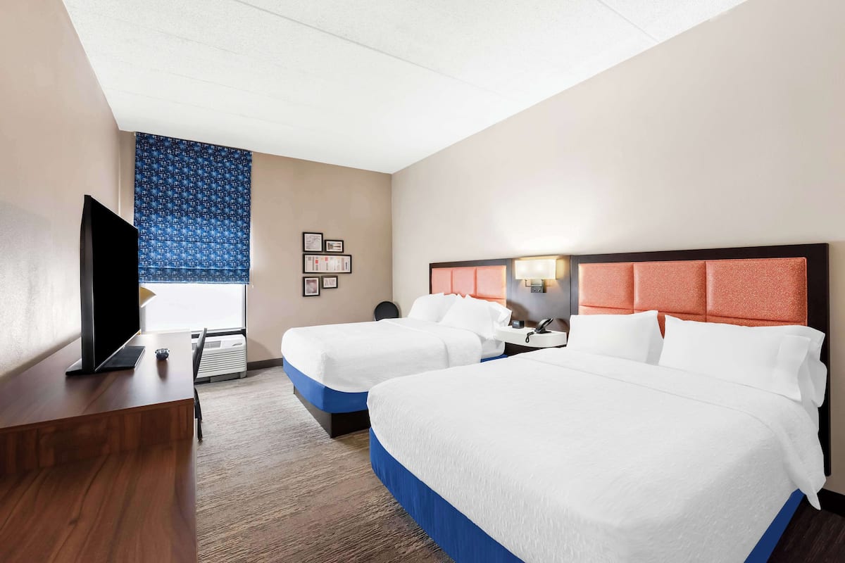 A hotel room with two beds, a TV, a desk, and a window with a patterned curtain.