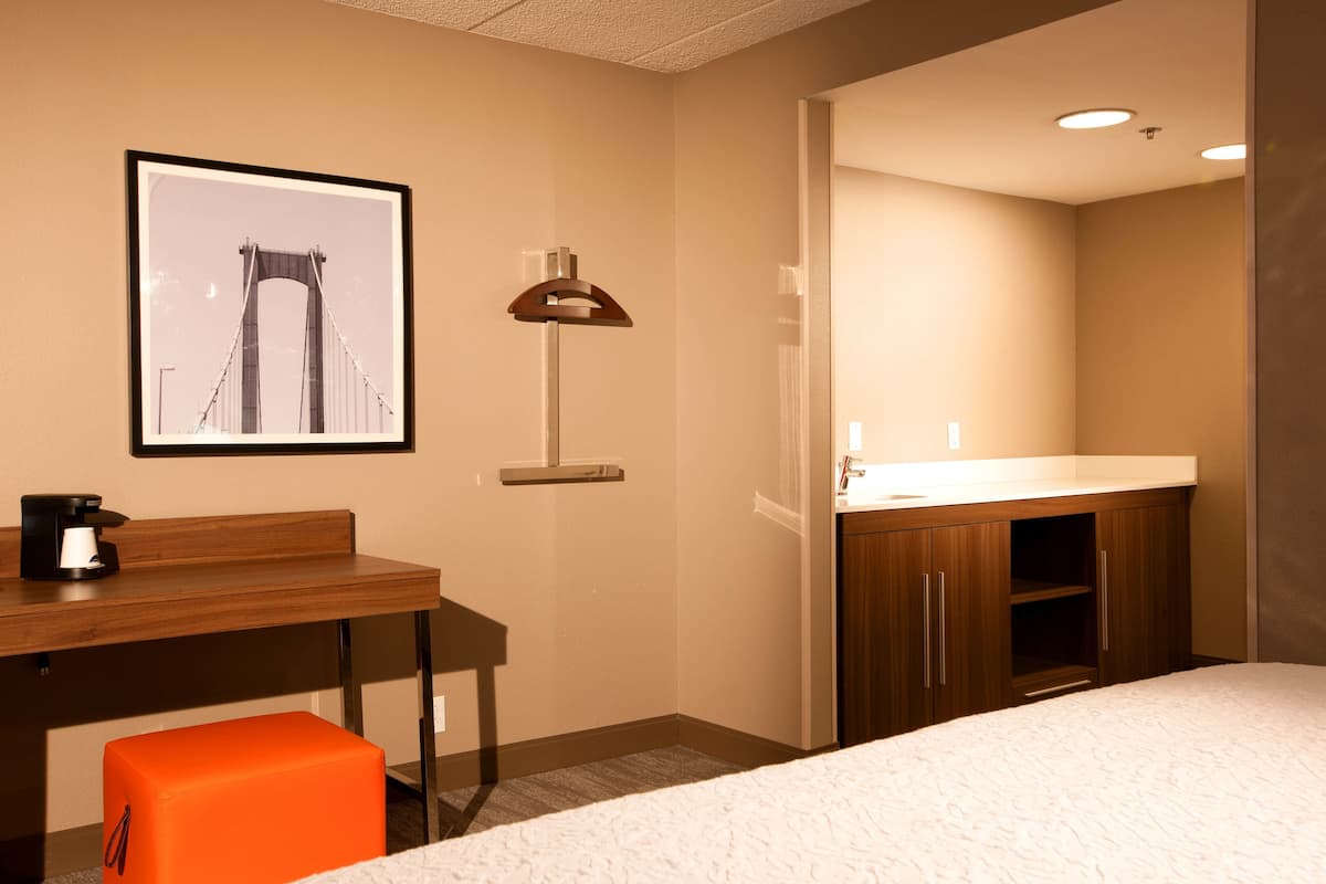 A hotel room with a bed, a desk with a coffee maker, a framed picture of a bridge, a lamp, and a bathroom with a sink and mirror.