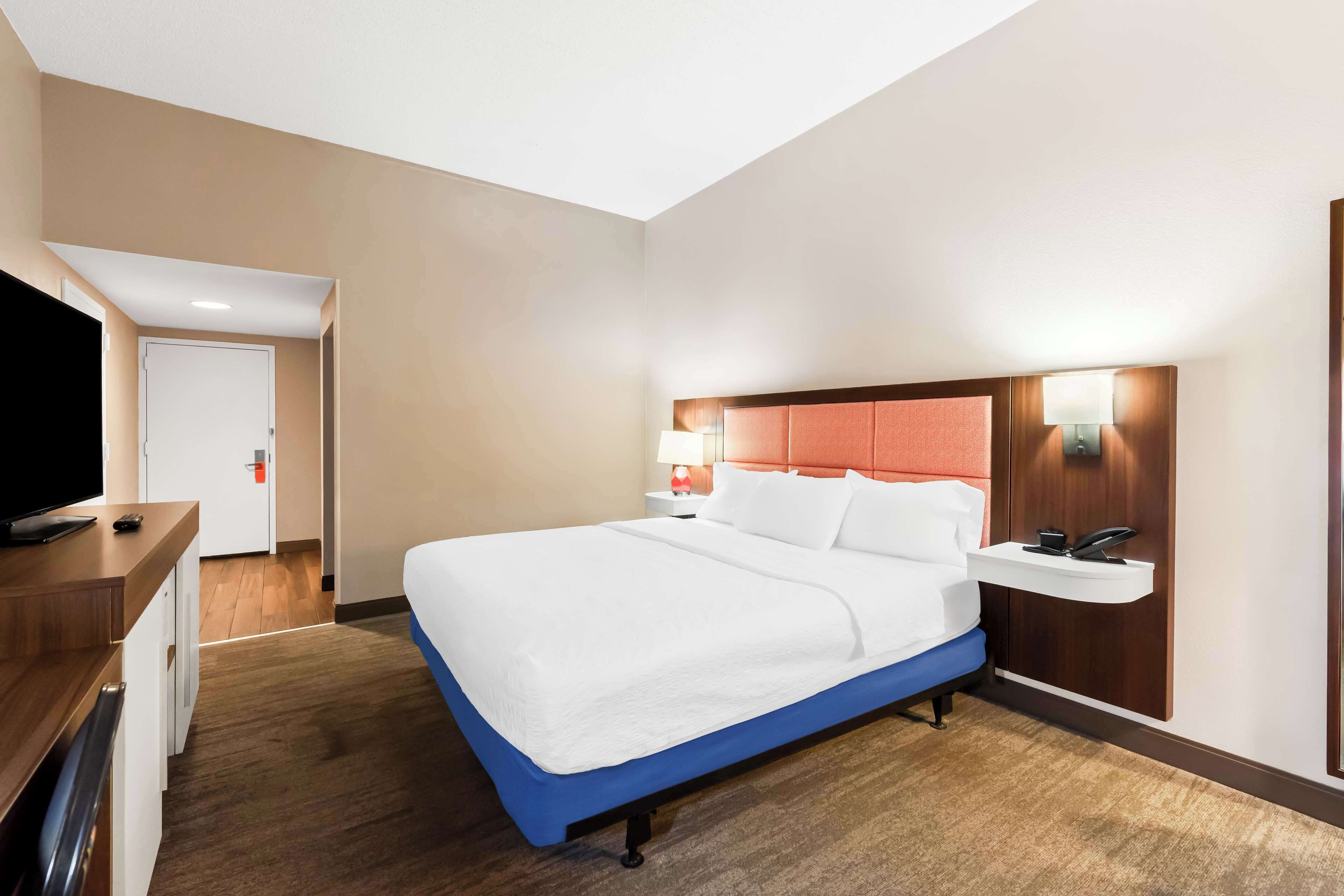 A hotel room with a bed, a desk, a TV, a nightstand, and a wall-mounted shelf.