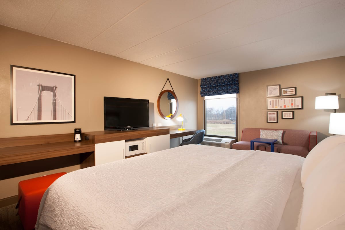 A hotel room with a large bed, a desk with a television, a chair, a small table, a window with a view, and a wall with framed pictures.