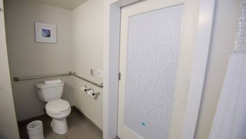 Un baño con inodoro, barra de apoyo, cubo de basura y puerta con diseño esmerilado.
