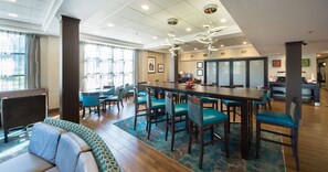 Una moderna hall d'hotel con un lungo tavolo condiviso, diverse poltrone blu, un divano e un tappeto decorato.