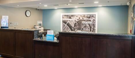 A hotel reception area with a clock, a bulletin board, a bulletin board with a travel advertisement, and a counter with a phone and a sign for Hilton Honors.