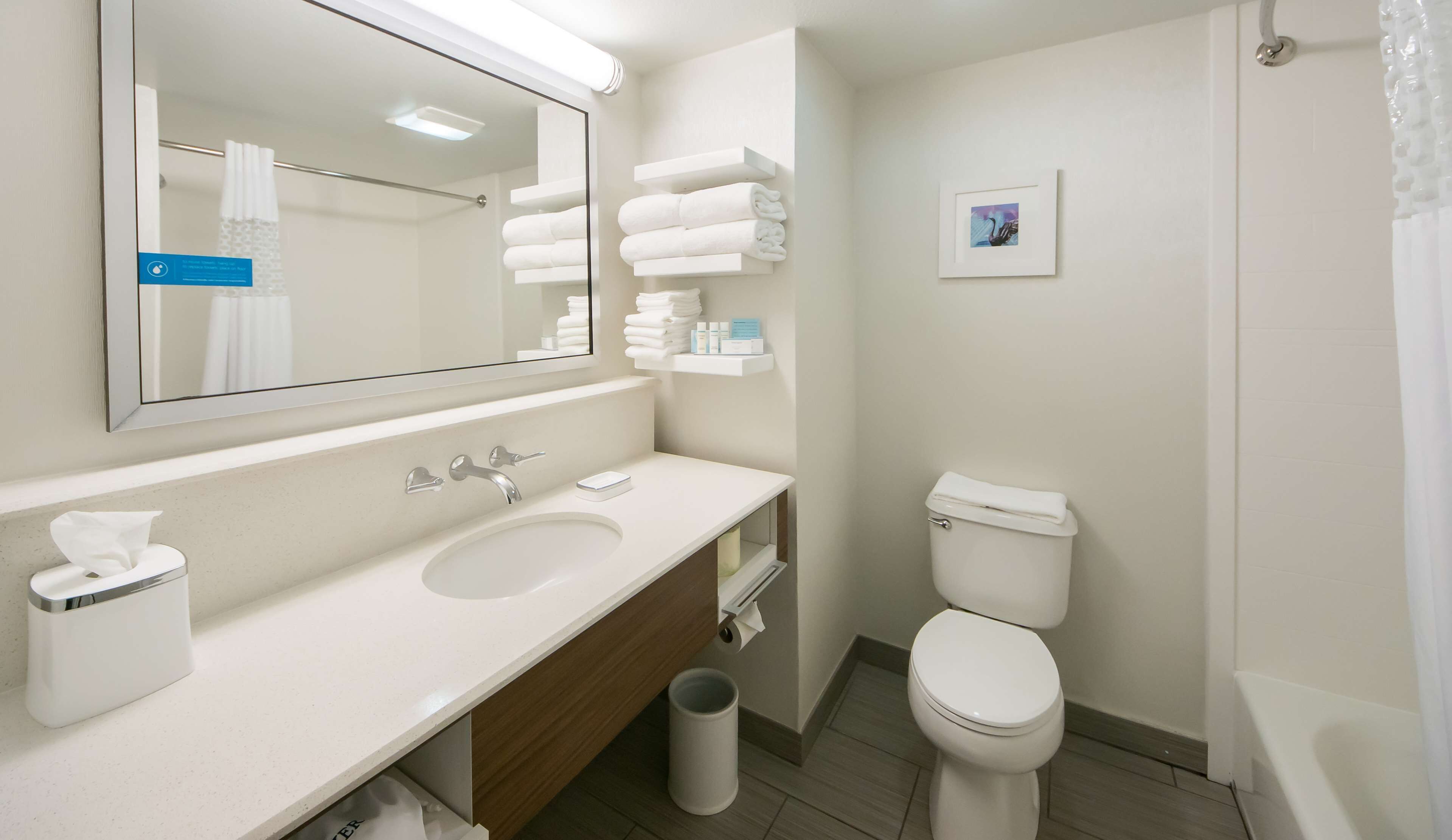 A modern bathroom with a white sink, mirror, and toilet. There are towels and toiletries on a shelf.