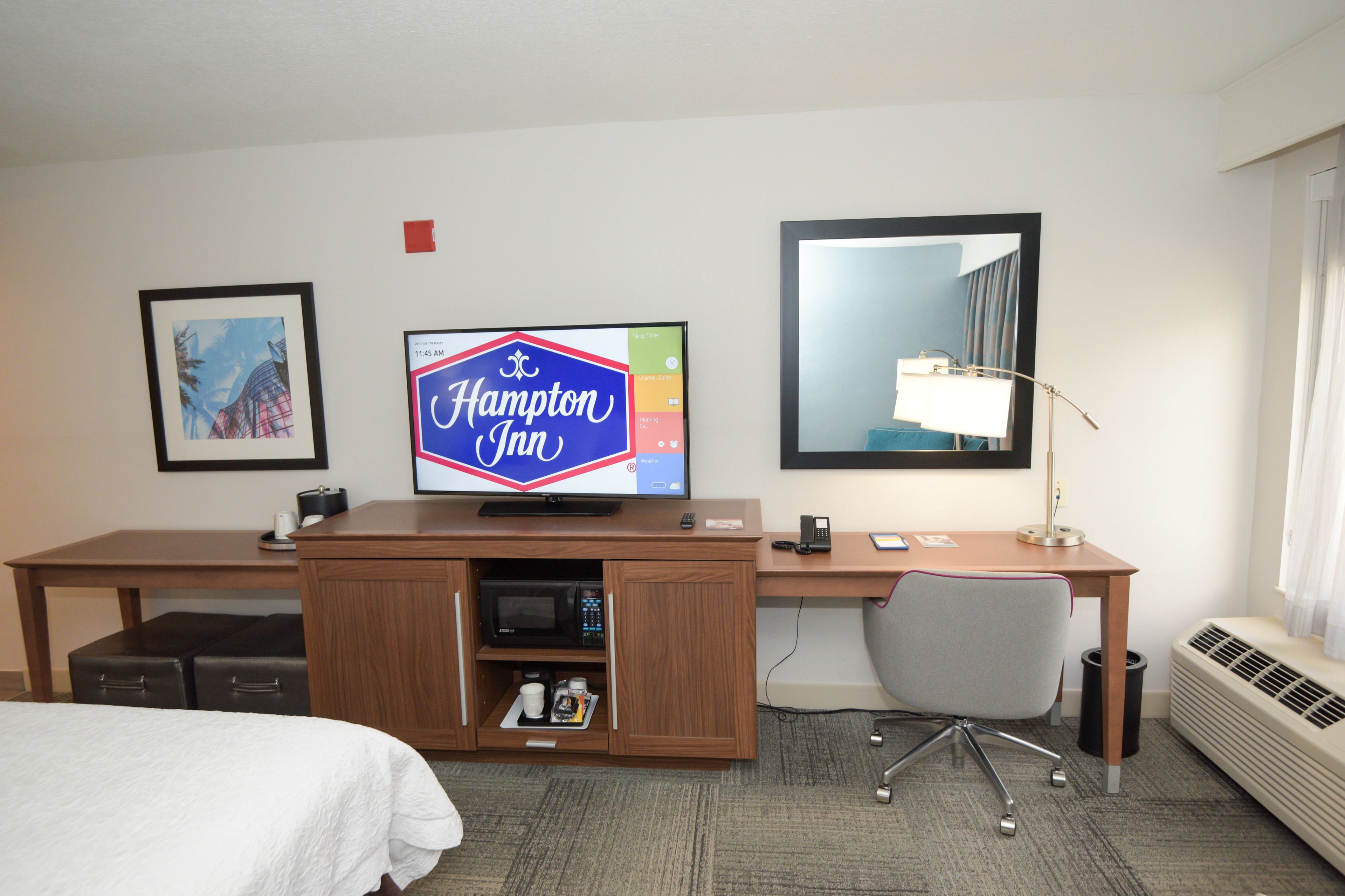 A hotel room with a bed, a desk with a computer, a television displaying 'Hampton Inn', a framed picture, and a large mirror.
