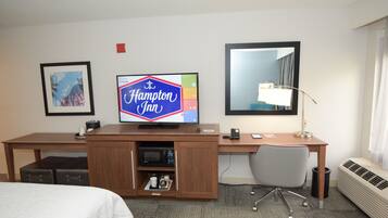A hotel room with a bed, a desk with a computer, a television displaying 'Hampton Inn', a framed picture, and a large mirror.