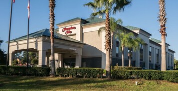 Un hotel Hampton Inn con fachada moderna, palmeras y un cielo azul despejado.