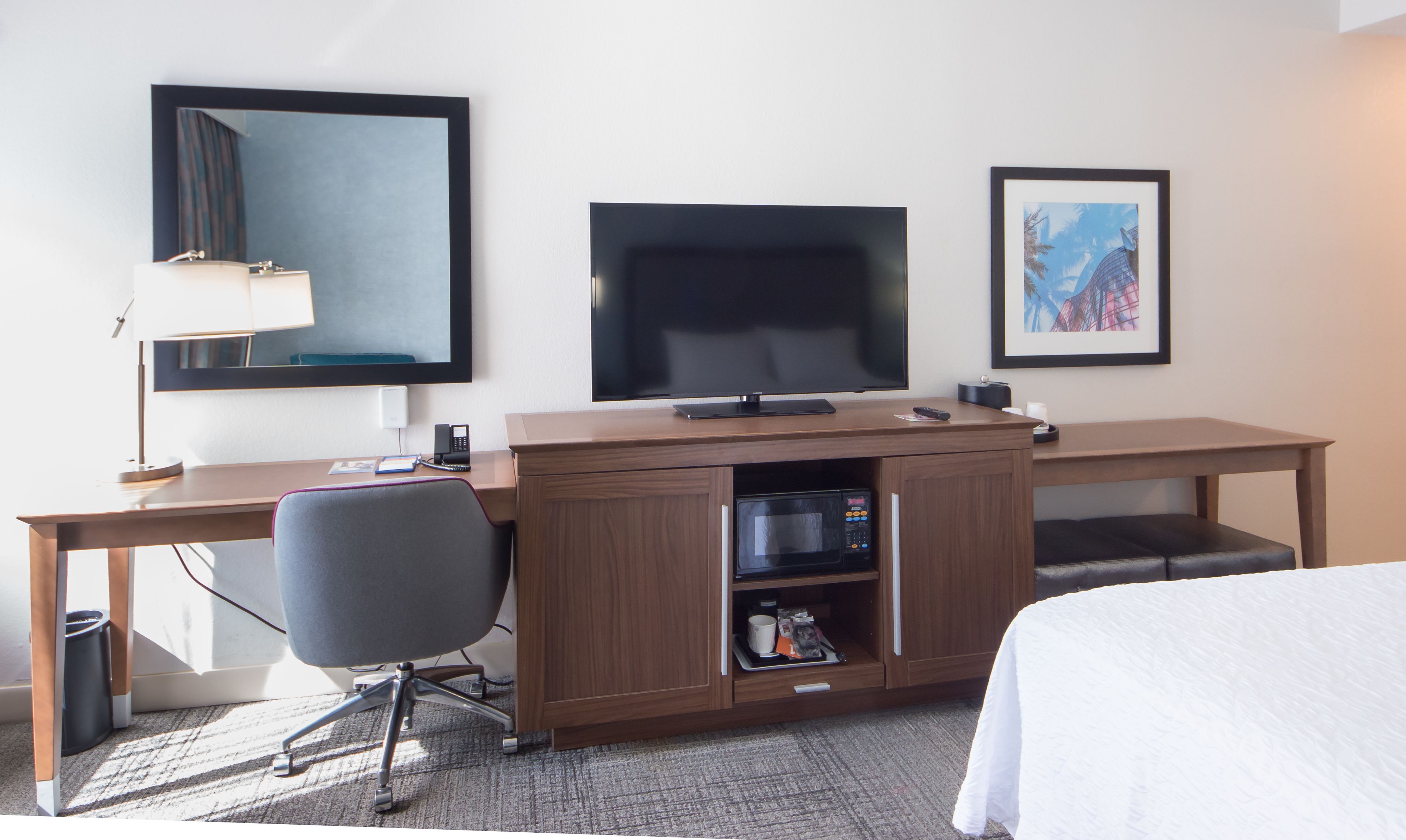 A hotel room with a wooden desk, a flat-screen TV, a telephone, a microwave, a chair, a bed, and a framed picture on the wall.