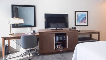 A hotel room with a wooden desk, a flat-screen TV, a telephone, a microwave, a chair, a bed, and a framed picture on the wall.