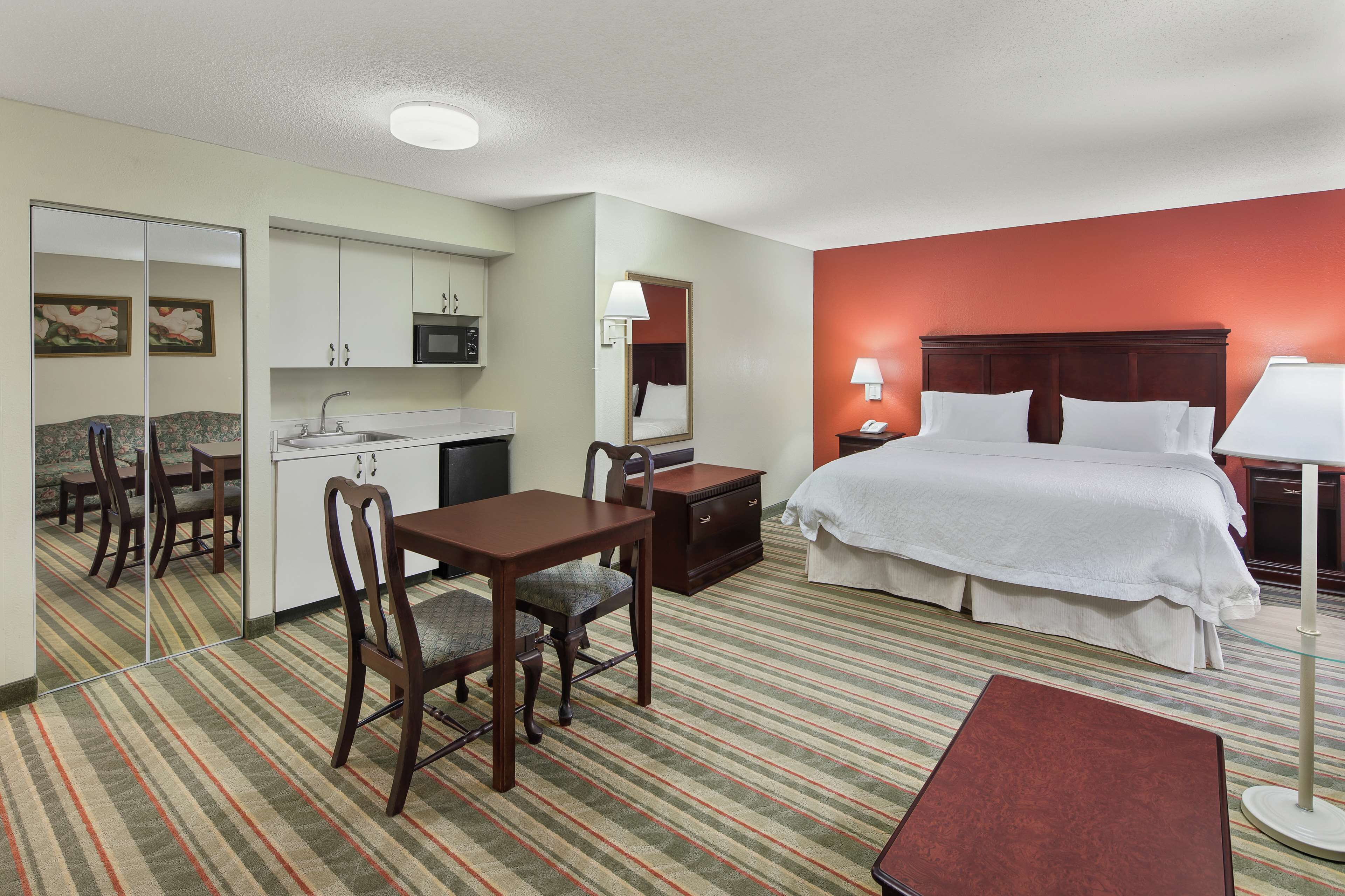 A hotel room with a bed, a desk with a chair, a small table, a mirror, a microwave, and a sink.